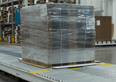 Shrink-wrapped unitized load of flat corrugated boxes moving on a conveyor belt.