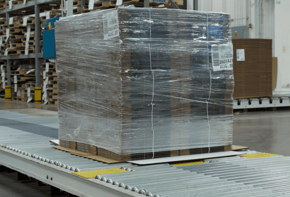 Shrink-wrapped unitized load of flat corrugated boxes moving on a conveyor belt.